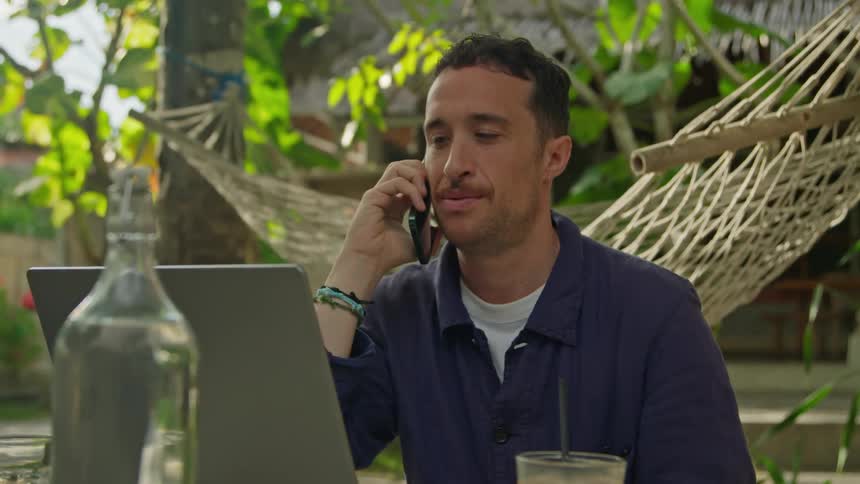A man is sitting outside at a table talking on his phone and typing on a laptop computer