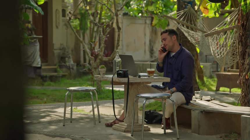 A man is sitting outside at a table talking on his phone and typing on a laptop computer