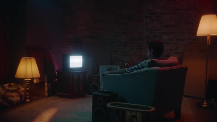 A young man sitting in an armchair shrugs his shoulders and starts to get up while the television plays static