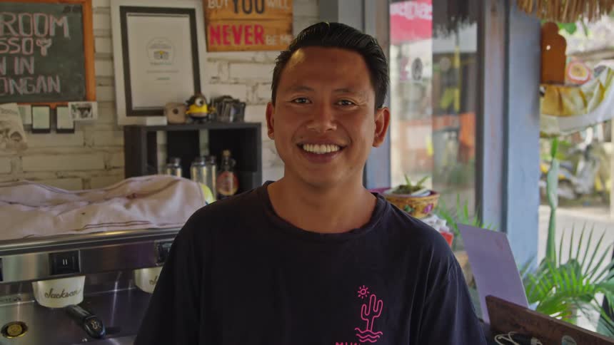 Proprietor of coffee shop stands smiling near counter open to the street