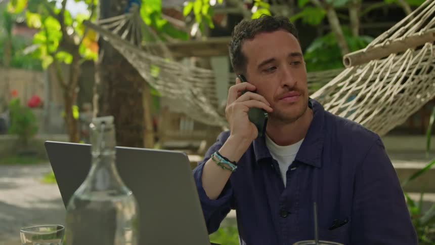 A man is sitting outside at a table talking on his phone and typing on a laptop computer