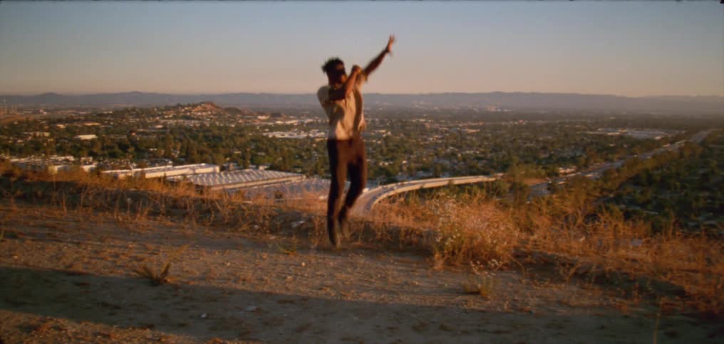 Young man wearing a white shirt dancing on a hill near a small town at evening