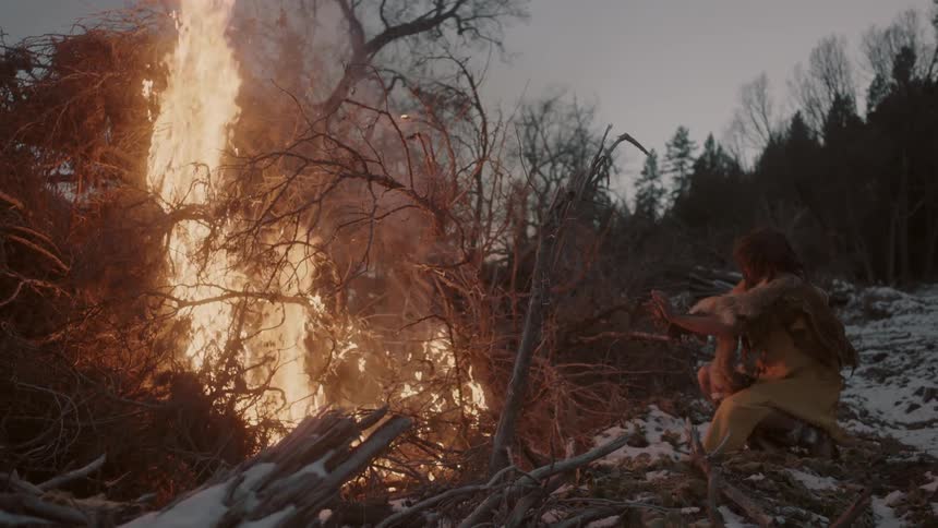 A man wearing furs and animal skins sits beside a bonfire on the snowy ground