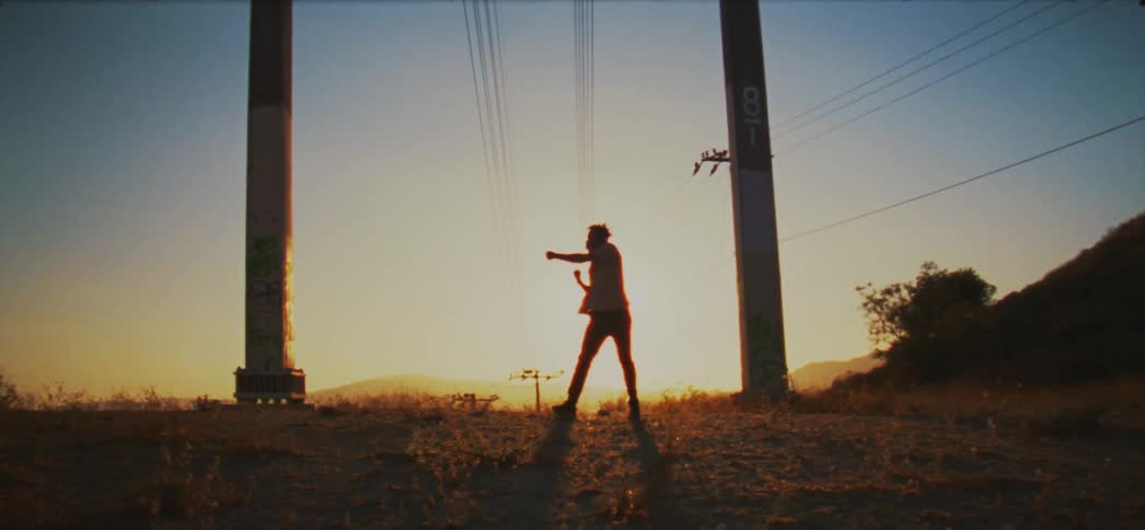 Silhouette of a man dancing between pillars with electrical wires hanging overhead and the sun shining brightly on the horizon