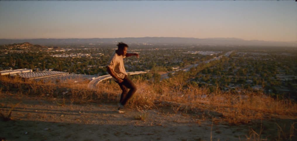 Young man wearing a white shirt dancing on a hill near a small town at evening