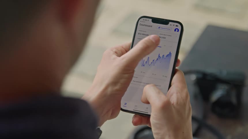 A man is holding his cell phone and using his thumb to move across a graph on the screen