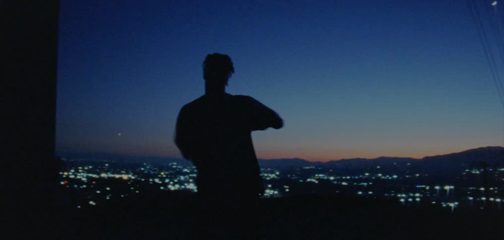 A man freestyle dances on a hill overlooking an urban suburb