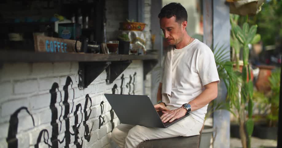 Man sitting on a stool on an outdoors coffee shop drinks while working on a laptop