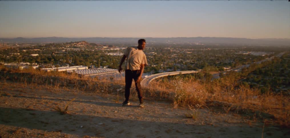 Young man wearing a white shirt dancing on a hill near a small town at evening