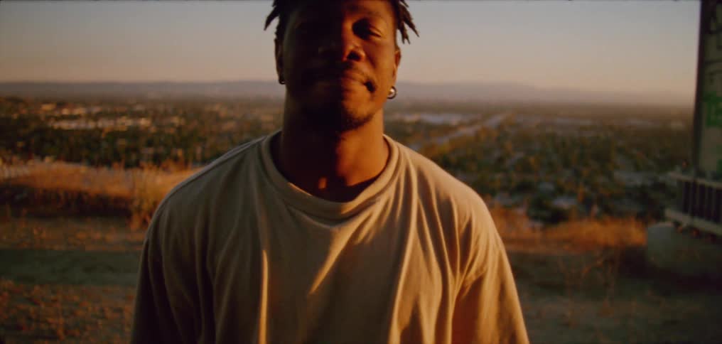 Sunlight shines on the face of a man standing outside with neighborhoods below in the distance under a clear sky