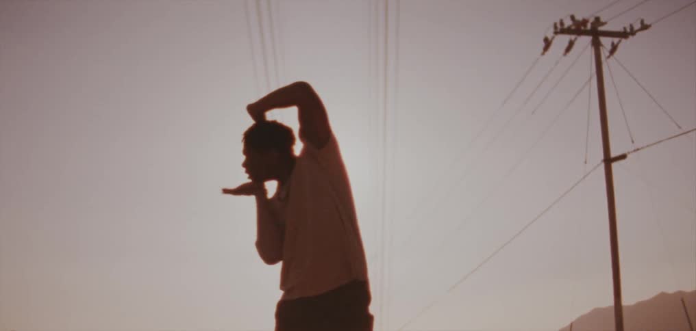 Man with dreadlocks and t shirt dancing is silhouetted against the sky in front of utility poles