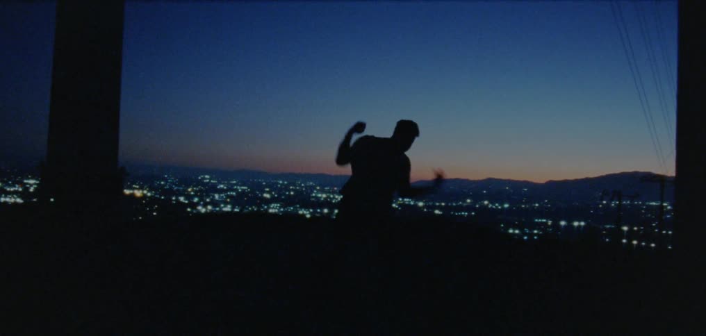 The silhouette of a young man dancing on a ridge above a valley with city lights glowing