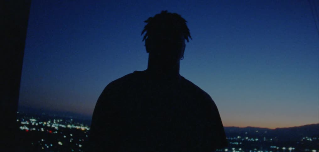 A man stands outside at dusk with the city skyline behind him and leans his head from side to side