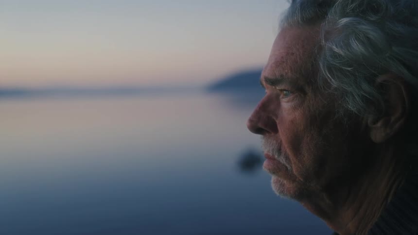 An elderly man with a mustache standing next to a calm lake