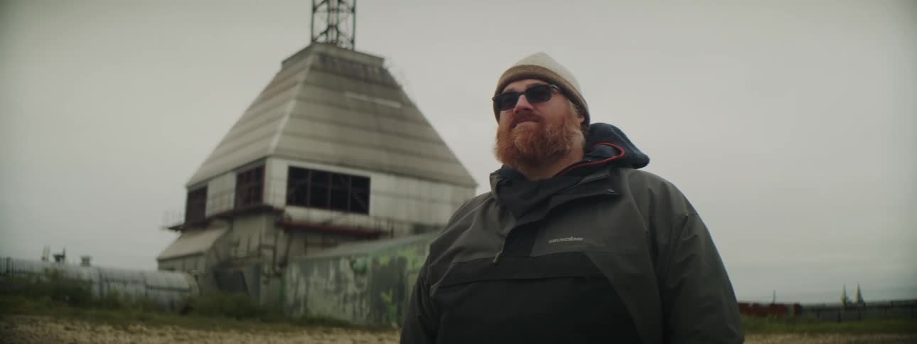 A man wearing a winter coat and hat with sunglasses is standing outside in front of a building