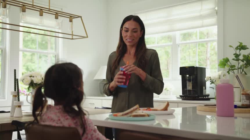 A woman tightens the lid of a straw cup and carries it over to a young girl eating at the kitchen counter