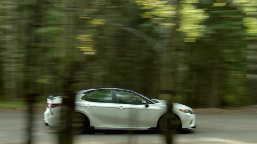 A white car travels on a road through the woods as a man standing near the trees watches it pass