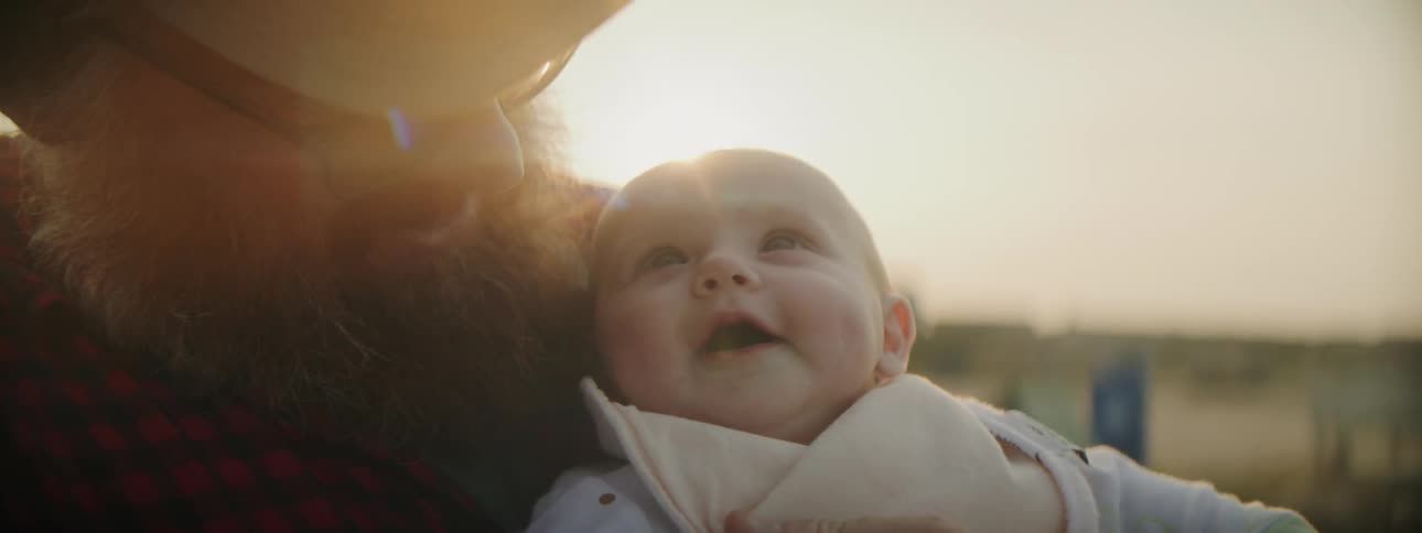 Man with beard and hat holds baby laughing in the field as the sun shines in the background