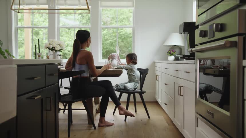Mother drinks a glass of water near her daughter playing with a toy over the kitchen table