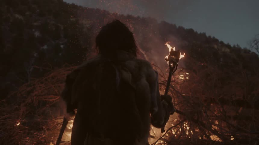 A man stands in front of a large fire holding a torch