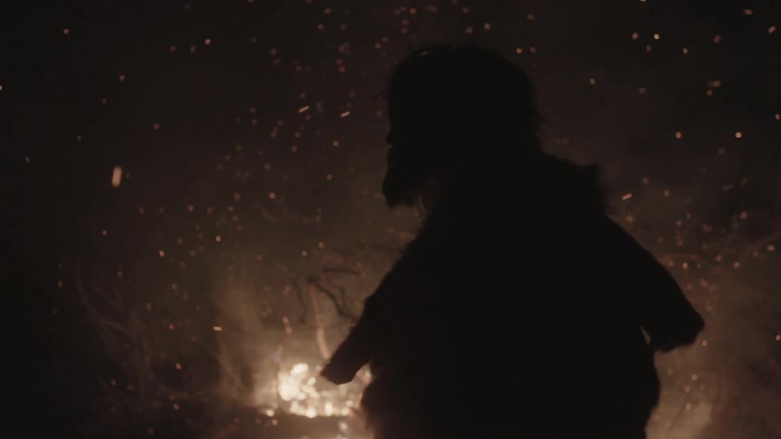 Bearded man in the wilderness walking in front of a fire