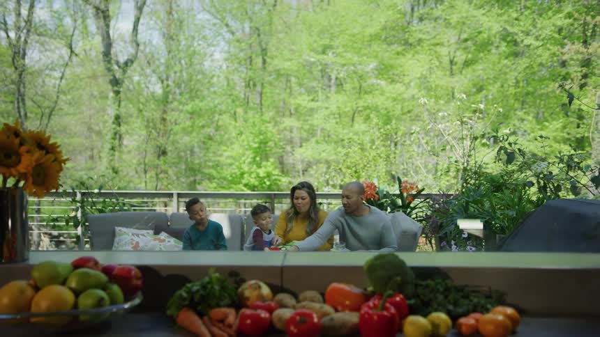 Family eats on patio while several vegetables and fruits sits clustered near window