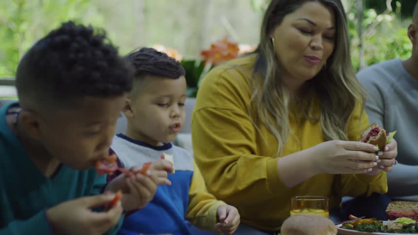 A little boy dances as he taps his burger sitting to eat outside with his family