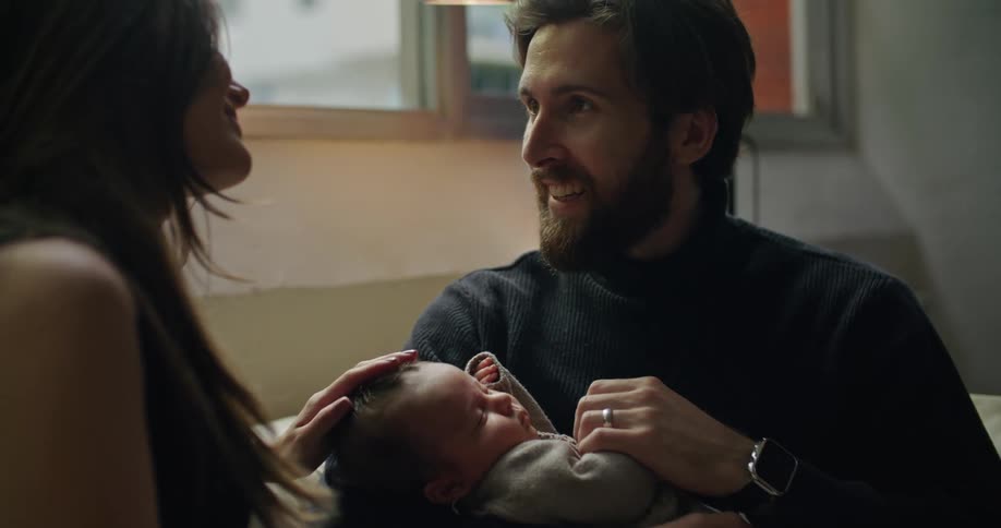Young happy couple talking while holding their newborn baby on the couch at home