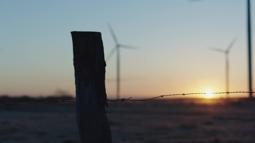 Wind turbines on a wind farm are on the other side of a barbed wire fence as the sun is on the horizon