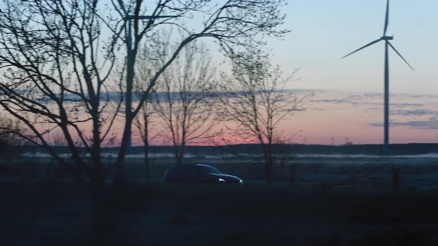 An automobile with the headlights on travels on a road near a lake and through a windfarm of wind turbines
