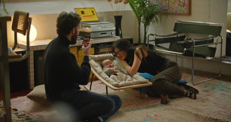 Young bearded man takes a picture of his couple posing next to their sleeping baby in the living room