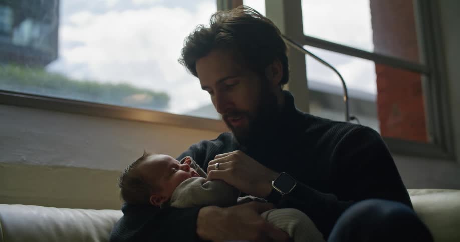 Bearded young father holding his newborn son on his arms sitting on a couch at home