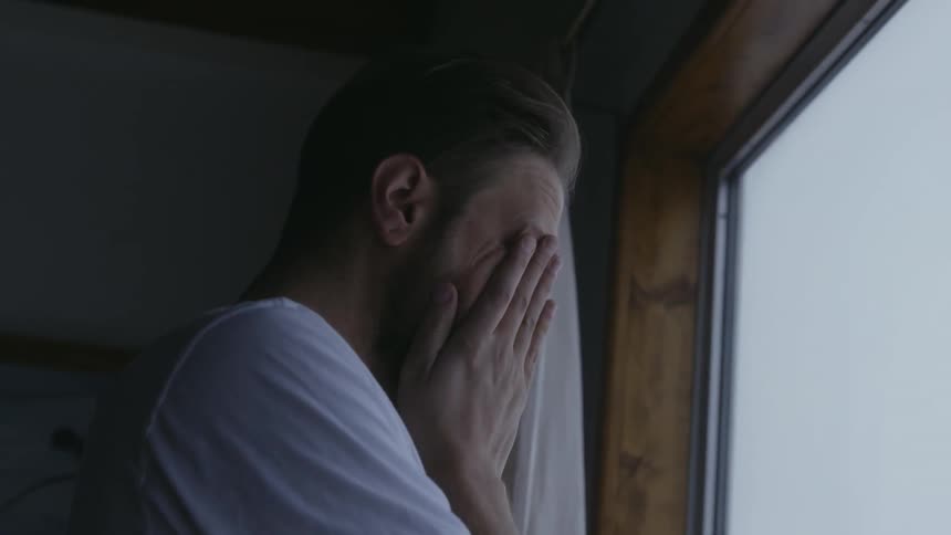 Man with beard and white shirt looks out the window rubbing his face in the morning
