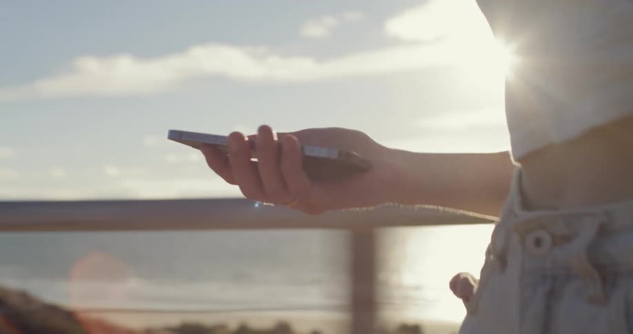 Young woman leisurely skateboards downhill with a phone on her hand next to the ocean