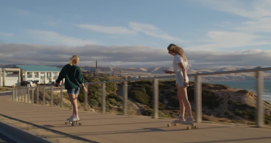 Two female skateboarders ride along sidewalk overlooking beach