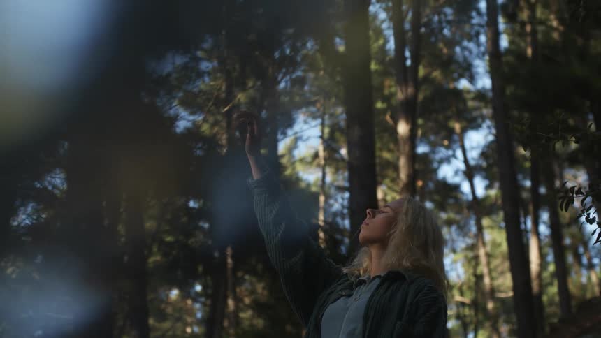 Woman stands in conifer forest reaching hand up toward the sky with her eyes closed