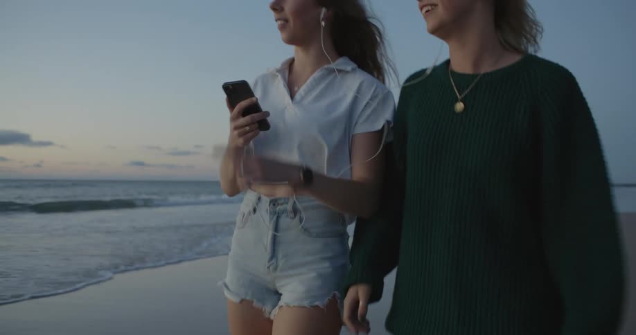 Two friends walking side by side on the beach each with an ear bud in smiling and laughing