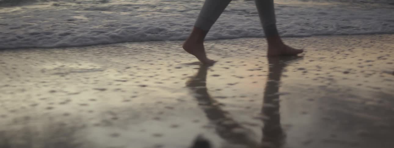 Following a barefoot woman walking on the wet sand on the coast of a beach at evening