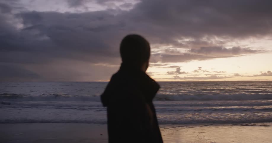 Man stands on beach looking out to red tinted horizon under dark clouds