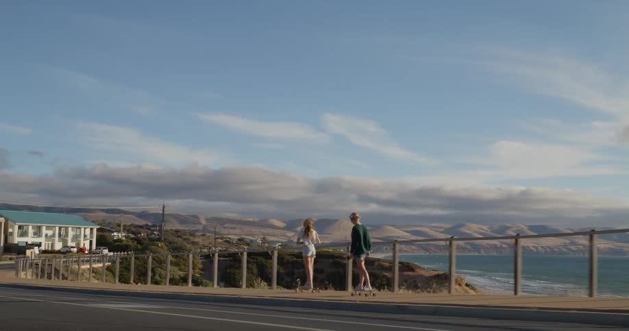 Two teenage girls are riding skateboards on a road overlooking the lake towards buildings