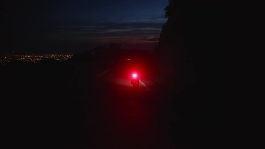 Cyclist rides bicycle with bright red taillight along highway overlooking cityscape at night