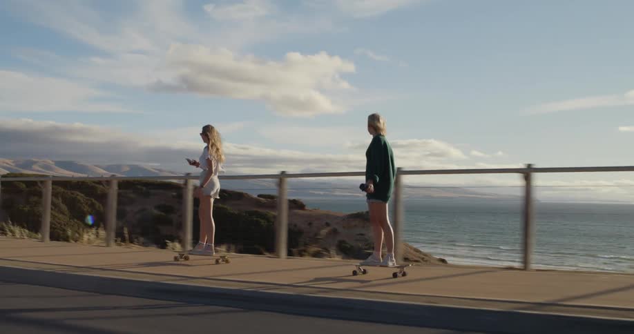 Two blonde women ride skateboards along sidewalk with promontory and ocean in the background
