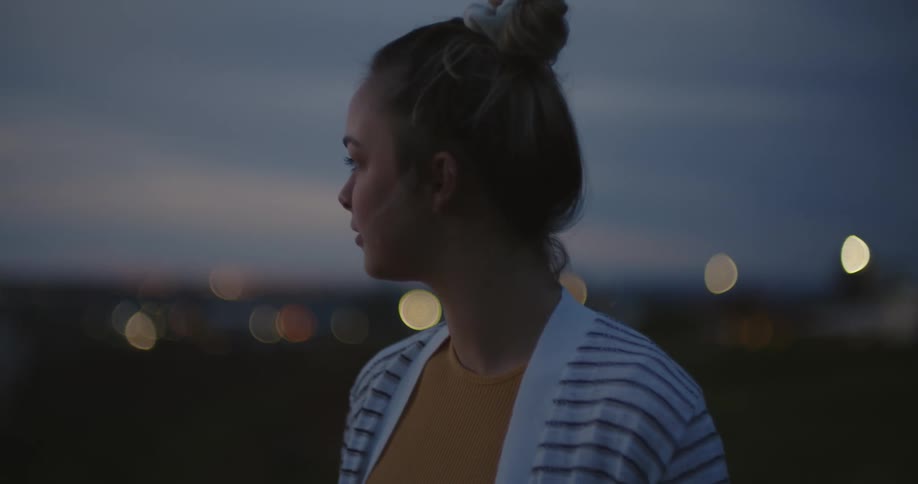 Young woman looks around atop a hill close to a distant waterfront town at evening