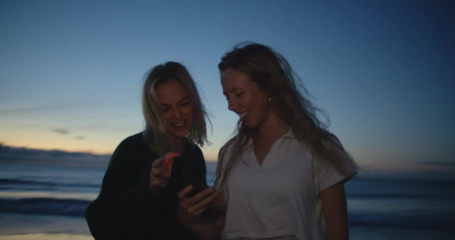 Two women laugh and look at a phone on the beach