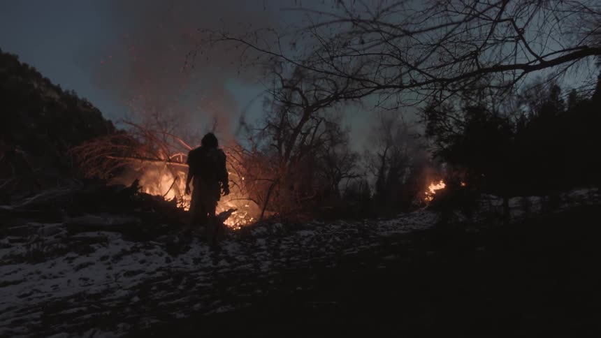 A man stands beside a brush pile burning on a hillside