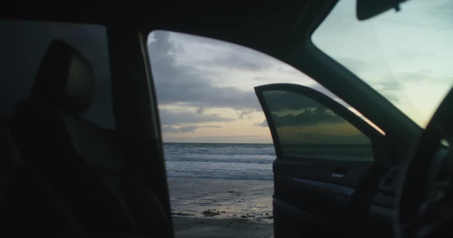 An automobile is parked at the beach and the door is open as waves roll into shore under a cloudy sky