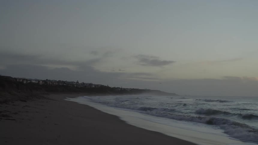 Rough water waves roll over the beach as seagulls fly into shore as dark clouds move over a town along the coast