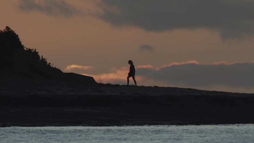 Silhouette of a woman climbing the gentle slope of a hill close to the coast at sunset