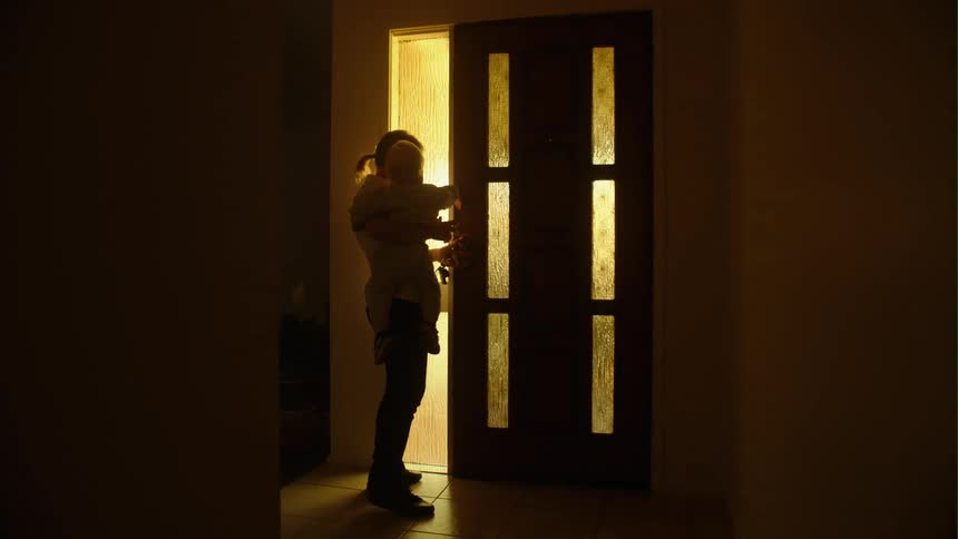 Mother with ponytail white shirt and jeans carries toddler son opening the door and puts him down in dark room