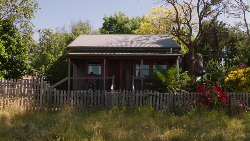An old man sits on the porch of a small house behind a picket fence and tall grass
